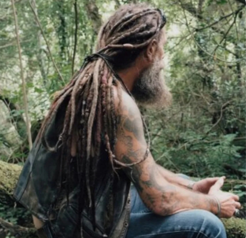 A man named Stewart with long, matted dreadlocks and a grey beard sits on a mossy log in a dense forest. He is wearing a leather vest and jeans, and his arms are covered in tattoos.